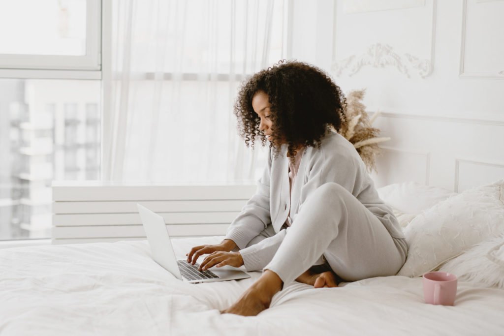 A Black woman in lounge wear sitting on a bed working on a laptop